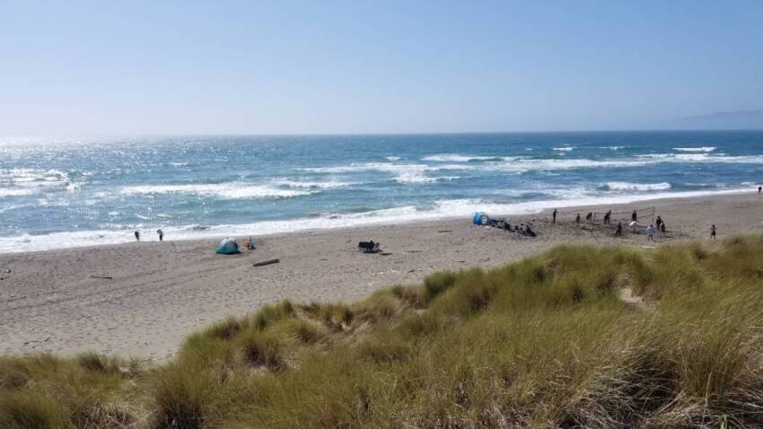 picture of blue ocean water with white foam waves and a few scattered people enjoying beach on the sand, taken from top of dune with green and brown grass in foreground